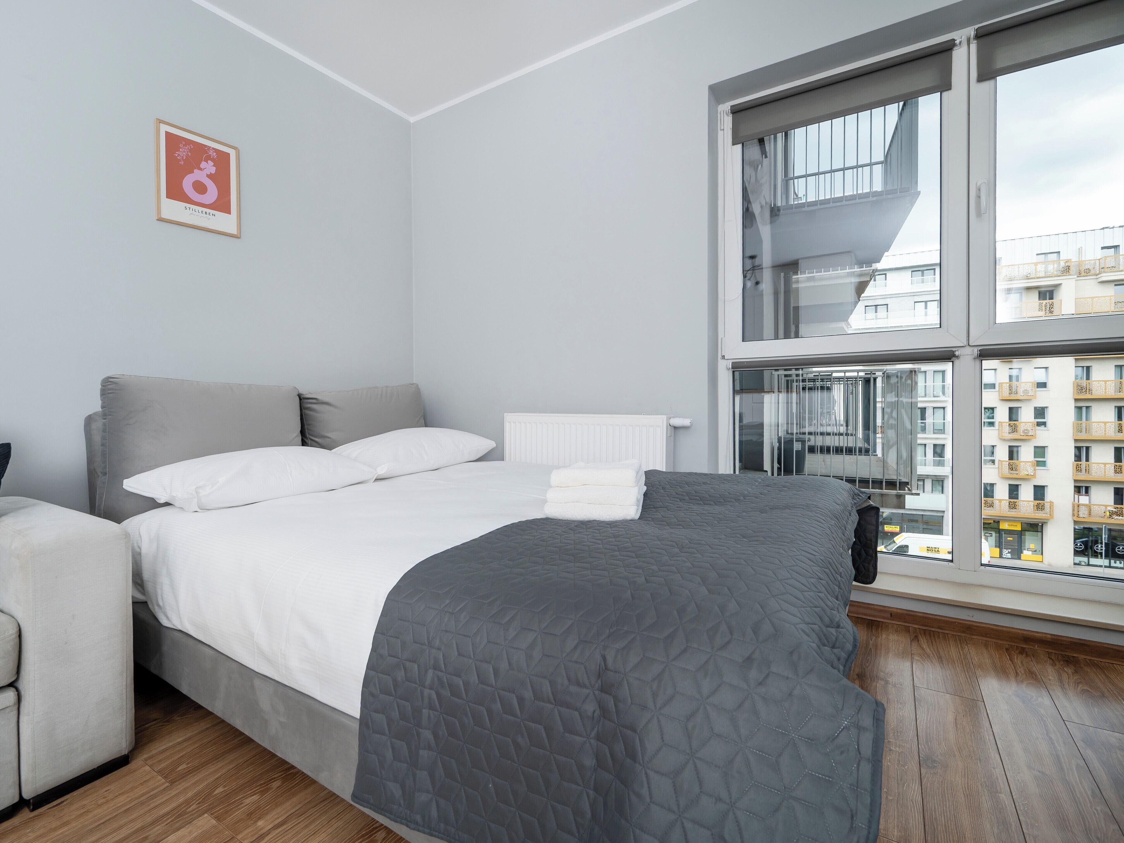 Een moderne slaapkamer met een bed, een radiator, een raam met uitzicht op gebouwen, en een lijst met een afbeelding aan de muur.