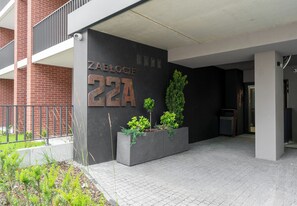Ein Gebäudeeingang mit einer schwarzen Wand, einem Metallschild mit der Aufschrift 'ZABLOCIE 22A' und einem Pflanzgefäß mit Pflanzen.