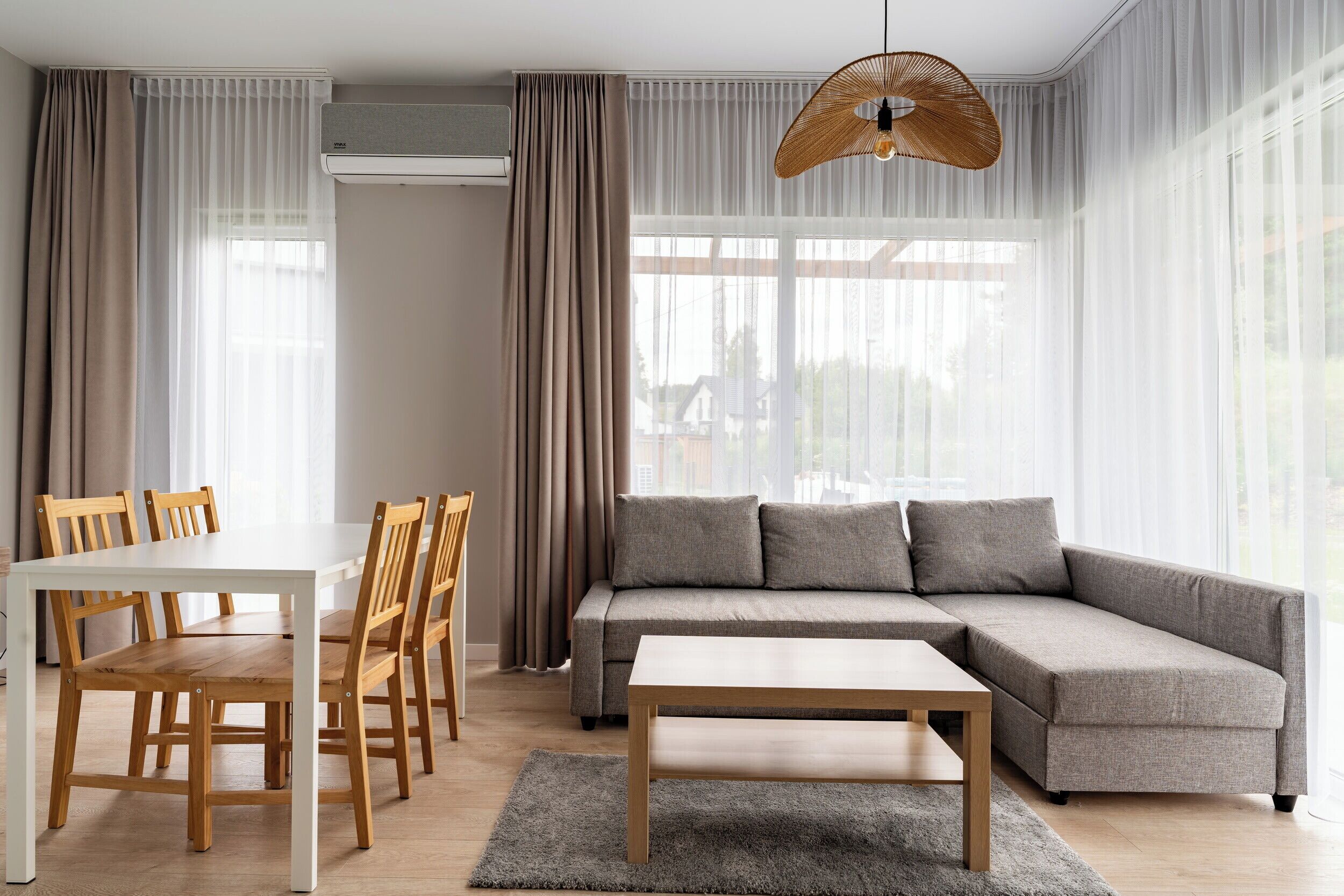 A modern living room with a sectional sofa, a dining table with chairs, and a large window with sheer curtains.