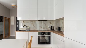 A modern kitchen with white cabinetry, a marble backsplash, a built-in oven, and a wooden dining table. - Willa 28D Lakeside Retreat Łączyno (Łączyno)