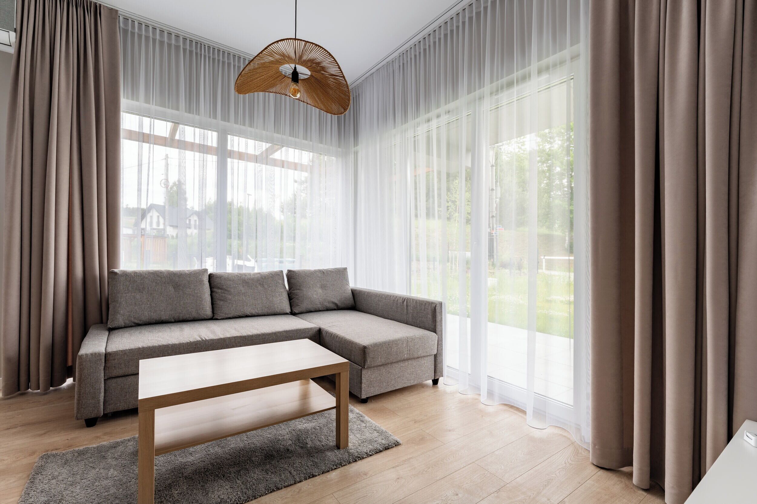 Un salon moderne avec un canapé d’angle, une table basse en bois et des rideaux légers.