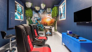 Een kamer met vier rode en zwarte gaming stoelen, een blauwe kast met een televisie, en een muurschildering met een fantasie thema.