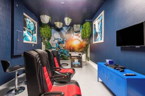 A room with four red and black gaming chairs, a blue cabinet with a television, and a wall mural with a fantasy theme. - Luxury 6BR Home with Private Pool (Davenport)