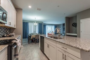 A modern kitchen with granite countertops, a dining area with a table and chairs, and a view of the living room through a glass door. - Luxury 6BR Home with Private Pool (Davenport)