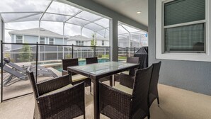 Een terras buiten met een glazen tafel en rieten stoelen, met uitzicht op een zwembad en omliggende huizen.