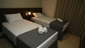 Zwei Einzelbetten mit weißen Bettwäsche und grauen Tagesdecken, ein kleiner Tisch mit einem Handtuch und eine Wandleuchte.