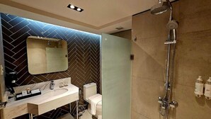A modern bathroom with a herringbone-patterned wall, a large mirror, a white sink, a toilet, and a shower with a handheld showerhead.