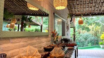 Disposição de buffet com luminárias de vime penduradas, piso de azulejos e vista para o verde lá fora.