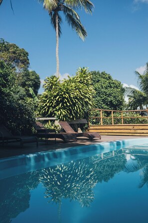Una piscina cristalina con el reflejo de palmeras y una terraza de madera con sillones de descanso.