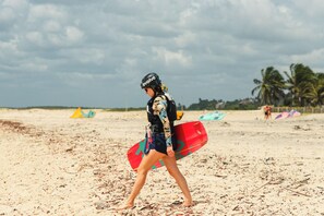 Una persona caminando sobre una playa arenosa con una tabla de surf, palmeras y sombrillas de playa al fondo.