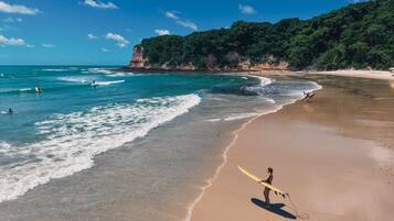 Praia com ondas, um surfista caminhando com uma prancha e um penhasco arborizado ao fundo.
