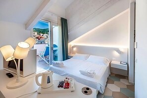 A hotel room with a bed, bedside table, lamp, and a view of the city. - Hotel Melograno (Spotorno)