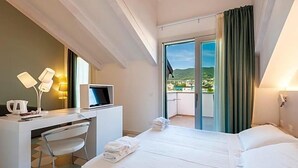 A hotel room with a bed, a desk with a computer, a chair, and a view of a balcony with a cityscape. - Hotel Melograno (Spotorno)