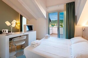A hotel room with a bed, a desk with a computer, a chair, and a view of a balcony with a cityscape. - Hotel Melograno (Spotorno)