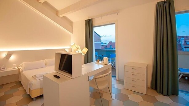 A hotel room with a bed, a desk with a TV, a chair, a dresser, and a window with curtains. - Hotel Melograno (Spotorno)