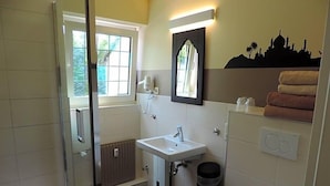 A bathroom with a glass shower enclosure, a white sink, a toilet, and a window with a view of greenery. - Hotel Sandmanns Am Dom (Cologne)