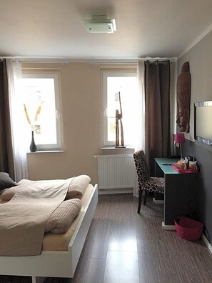 A bedroom with a bed, a desk, a chair, a radiator, and a window with curtains. - Hotel Sandmanns Am Dom (Cologne)