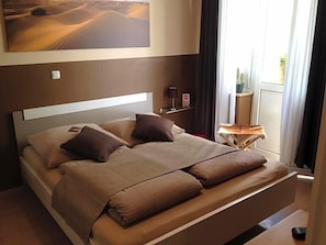 A bed with a headboard, pillows, and a blanket. There is a small table with a lamp and a plant. - Hotel Sandmanns Am Dom (Cologne)