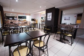 A restaurant with multiple tables and chairs, a kitchen area, and a clock on the wall. - Hã´tel Akena Nantes (Sainte-Luce-sur-Loire)