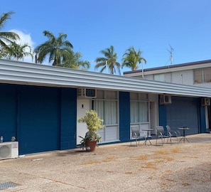 A motel with a covered outdoor seating area, air conditioning units, and a potted plant.