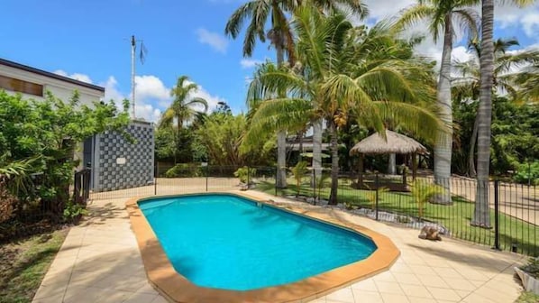 A swimming pool with a tiled deck, surrounded by palm trees and a fenced area.