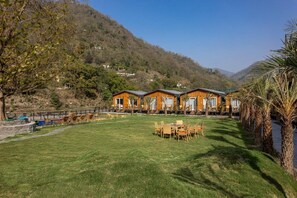 A row of wooden cabins with a grassy lawn and palm trees in the foreground. - Justa Rasa Retreat & Spa Rishikesh (Yamkeshwar)