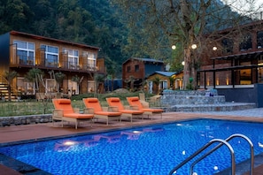 A swimming pool with lounge chairs, a stone wall, and buildings in the background. - Justa Rasa Retreat & Spa Rishikesh (Yamkeshwar)