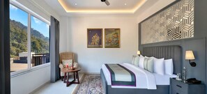 A bedroom with a large bed, a chair, a nightstand, and a view of a mountainous landscape. - Justa Rasa Retreat & Spa Rishikesh (Yamkeshwar)