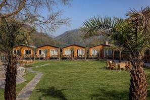 A row of wooden cabins with outdoor seating, surrounded by a grassy lawn and palm trees, set against a mountainous backdrop. - Justa Rasa Retreat & Spa Rishikesh (Yamkeshwar)