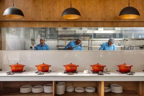 A kitchen with chefs preparing food, a buffet table with orange pots, and a stack of plates. - Justa Rasa Retreat & Spa Rishikesh (Yamkeshwar)