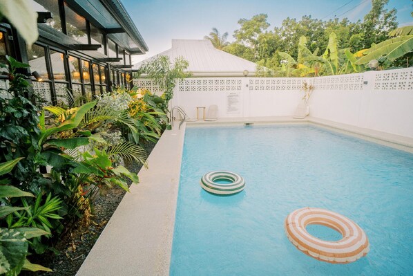 Una piscina exterior amb dos anells, envoltada de vegetació abundant i una tanca blanca.