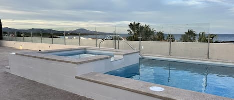A rooftop pool with two hot tubs, a glass balustrade, and a view of the sea and mountains.