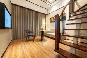 A hotel room with a wooden floor, a sofa, a chair, a small table with a lamp, and a TV.