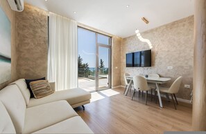 A modern living room with a sofa, a dining area with a table and chairs, a flat-screen TV mounted on the wall, and a large sliding glass door leading to an outdoor area. - Sapphire Coast Drymades Modern 2BR Apartment (Himarë)
