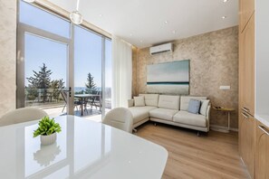 Ein modernes Wohnzimmer mit Glastür zum Balkon, ein weißes Sofa, ein Esstisch mit Pflanze und ein Gemälde an der Wand.