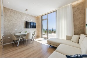 Ein modernes Wohnzimmer mit einer Couch, einer Essecke mit Stühlen und Tisch, einem an der Wand befestigten Flachbildfernseher und einer großen Schiebeglastür, die auf einen Balkon mit Blick freigibt.