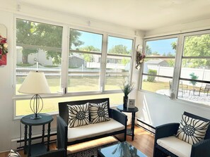 A sunlit room with a sofa, chairs, a table, and large windows offering a view of a fenced backyard.