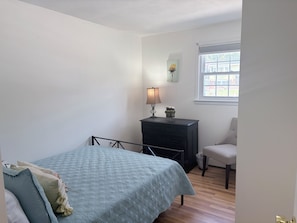 Un dormitorio con cama, mesita de noche, lámpara, silla y ventana.