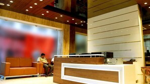 A reception area with a wooden reception desk, a waiting area with a sofa, and a large window. - Mango Hotels Vybrant Vijayawada by Othpl (Vijayawada)