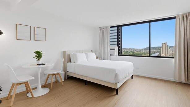 Ein modernes Hotelzimmer mit einem großen Bett, einem kleinen Tisch mit Stühlen, einer Zimmerpflanze und einem großen Fenster mit Blick auf die Stadt.