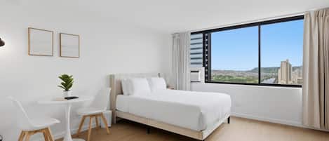 A modern hotel room with a large bed, a small table with chairs, a potted plant, and a large window offering a city view.