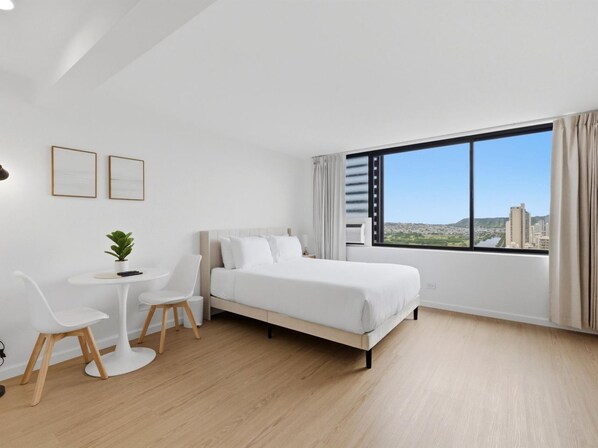 Habitación de hotel moderna con una cama grande, una mesita con sillas, una planta en maceta y un amplio ventanal con vistas a la ciudad.