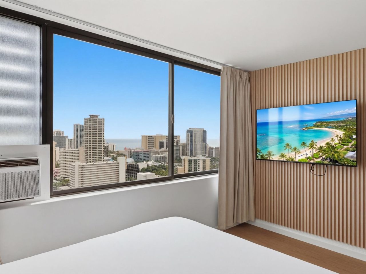 A hotel room with a large window offering a cityscape view, a flat-screen TV mounted on the wall, and a white bed.