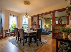 A dining area with a wooden table and chairs, a chandelier, and a view into another room with a green wall and a clock.