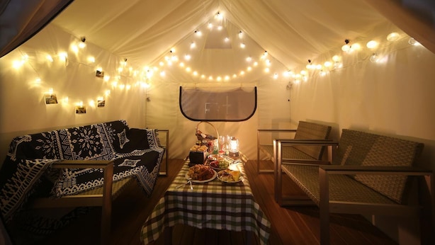 Un espacio amueblado con tienda de campaña, mantel a cuadros, dos sillones cómodos, una pequeña mesa con comida y bebidas, y luces colgantes.