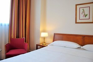 Una habitación de hotel con cama, una mesita de noche con lámpara, una silla roja y un cuadro en la pared.