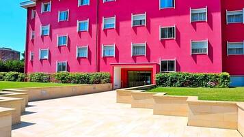 Un edificio de varios pisos con fachada roja, una entrada central y un patio ajardinado con escalones que conducen a la entrada.
