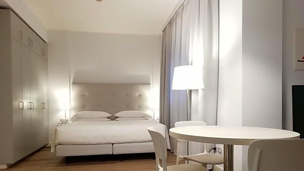 A bedroom with a bed, bedside table, lamp, and a dining area with chairs. - Delta Hotel (San Donato Milanese)