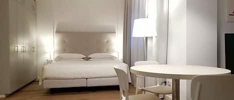A bedroom with a bed, bedside table, lamp, and a dining area with chairs.