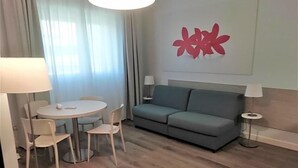 A modern living room with a grey sofa, a round dining table with chairs, and a wall-mounted lamp. - Delta Hotel (San Donato Milanese)